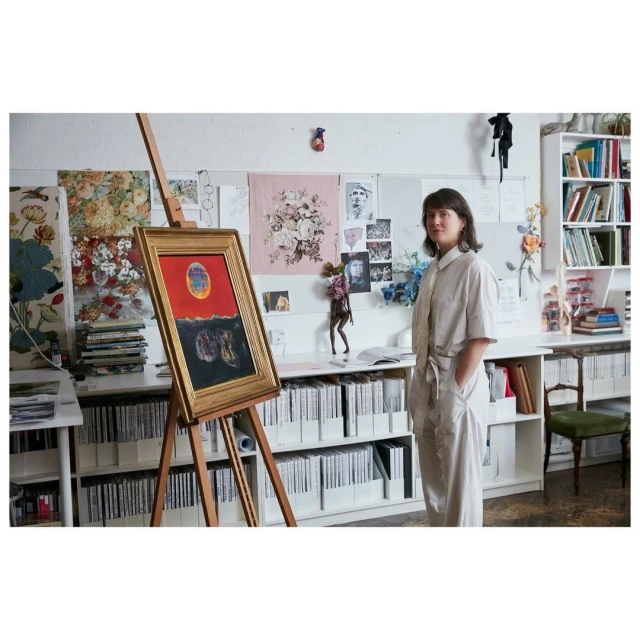 An artist stands next to a wood easel displaying her painting with a red and black color palette inside an art studio.