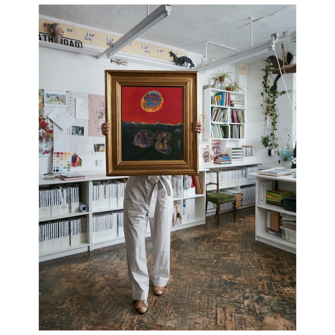 Person stands in an office, holding a framed painting of a red sky with dark land masses, wearing tan shoes and white pants.