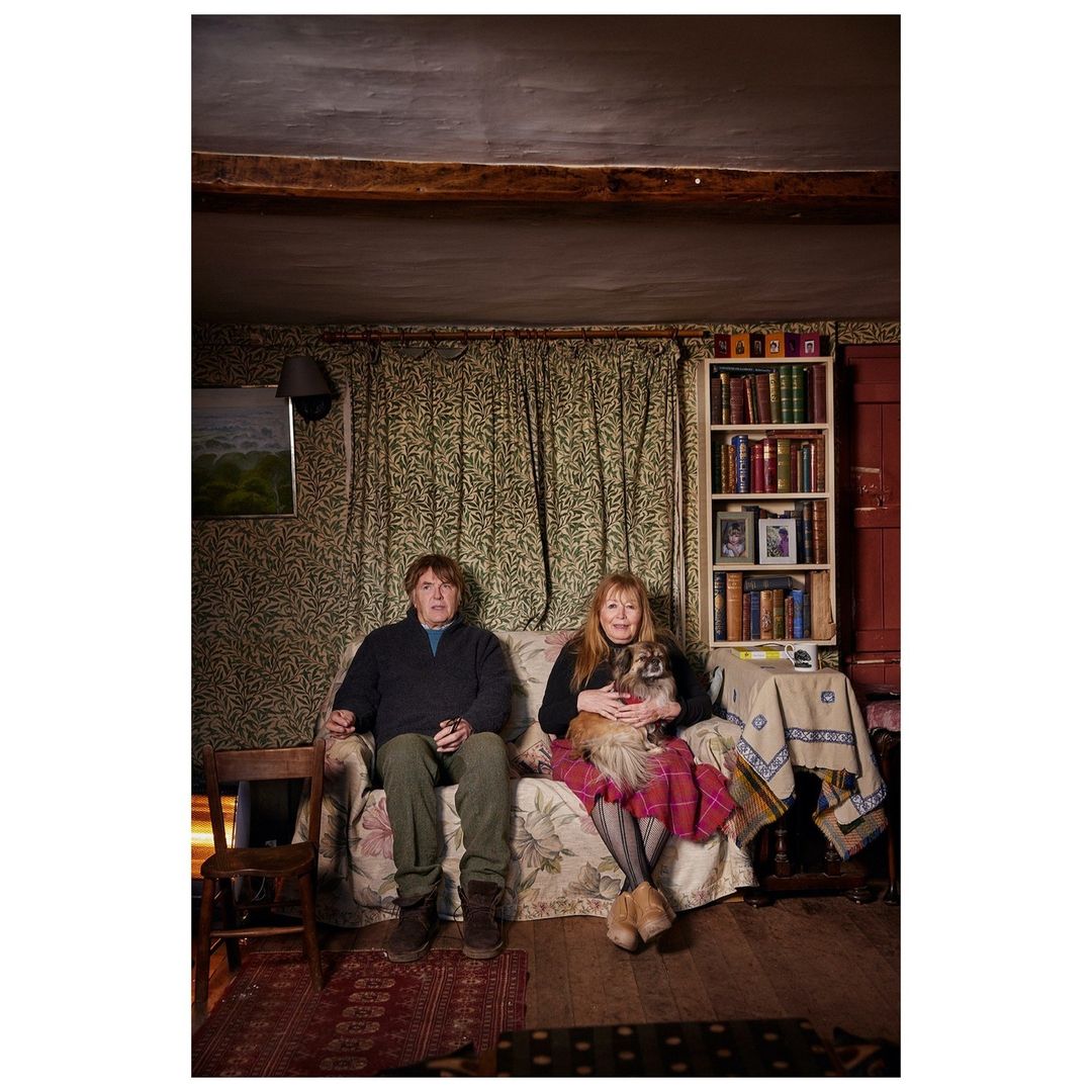 Mature couple sits on a floral sofa in a rustic living room with their dog. Bookshelves, patterned wallpaper, and a wooden chair are visible.