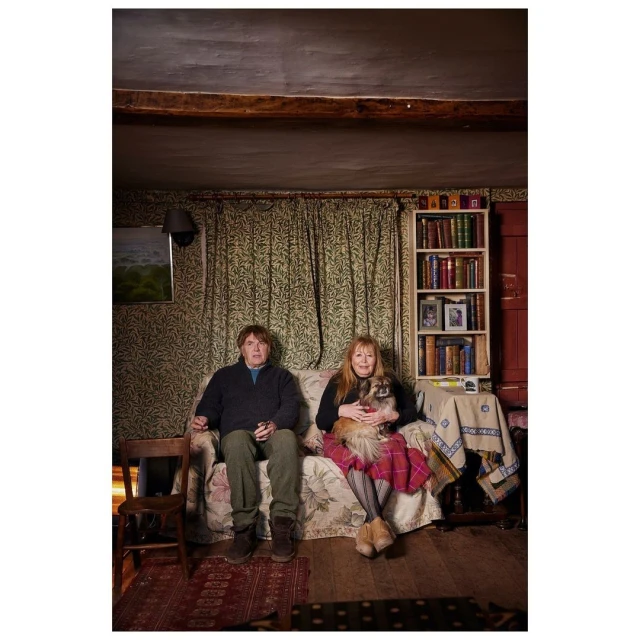 Mature couple sits on a floral sofa in a rustic living room with their dog. Bookshelves, patterned wallpaper, and a wooden chair are visible.