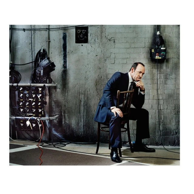 Adult man in a suit sits on a wooden chair in front of a weathered brick wall with lighting equipment and electrical box.
