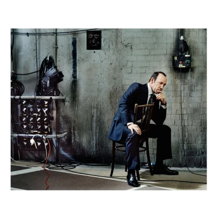 Adult man in a suit sits on a wooden chair in front of a weathered brick wall with lighting equipment and electrical box.