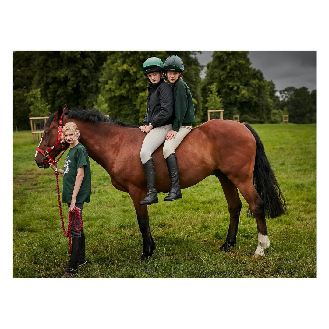 Two girls with riding helmets sit on a brown horse in a field, while another girl holds the horse's reins outdoors during the daytime.