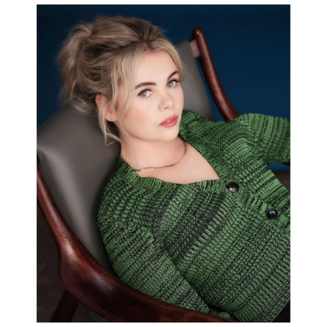 Blonde woman in a green knit sweater is sitting in a dark wooden chair against a dark blue background in a portrait-style image.