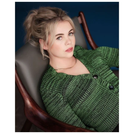Blonde woman in a green knit sweater is sitting in a dark wooden chair against a dark blue background in a portrait-style image.