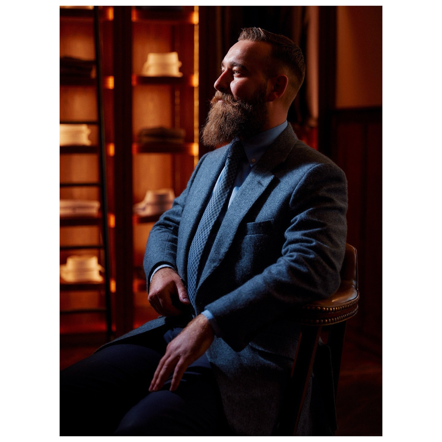 Bearded adult male in tweed suit and tie poses, sitting in a dimly lit room with shelves holding folded shirts and hats.