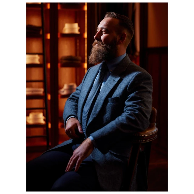 Bearded adult male in tweed suit and tie poses, sitting in a dimly lit room with shelves holding folded shirts and hats.