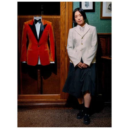 Portrait of a woman in a blazer posing next to an orange tuxedo on a mannequin in an indoor setting with patterned wallpaper.