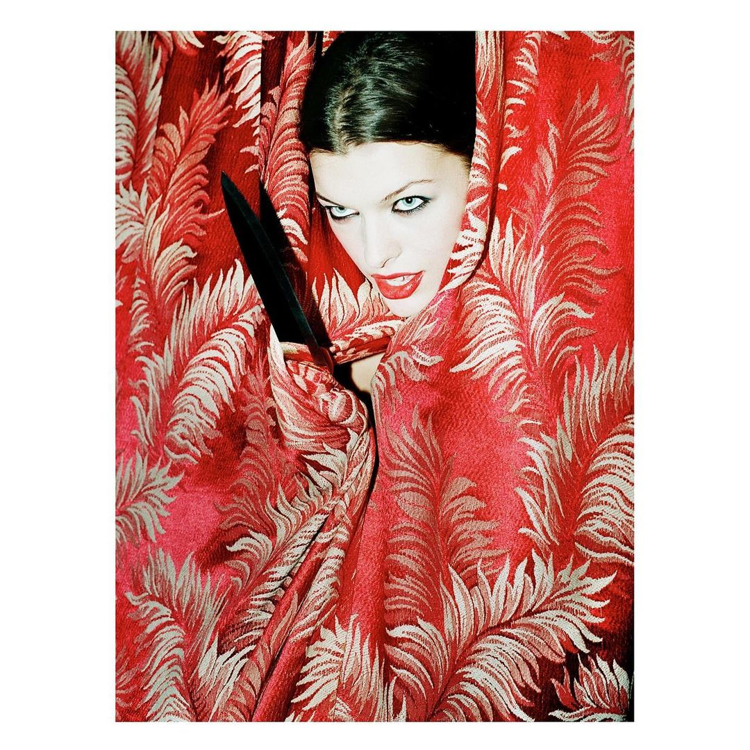 Close-up of a woman with fair skin, dark hair, and red lipstick. She is wrapped in red floral fabric, with a black object in her hand.