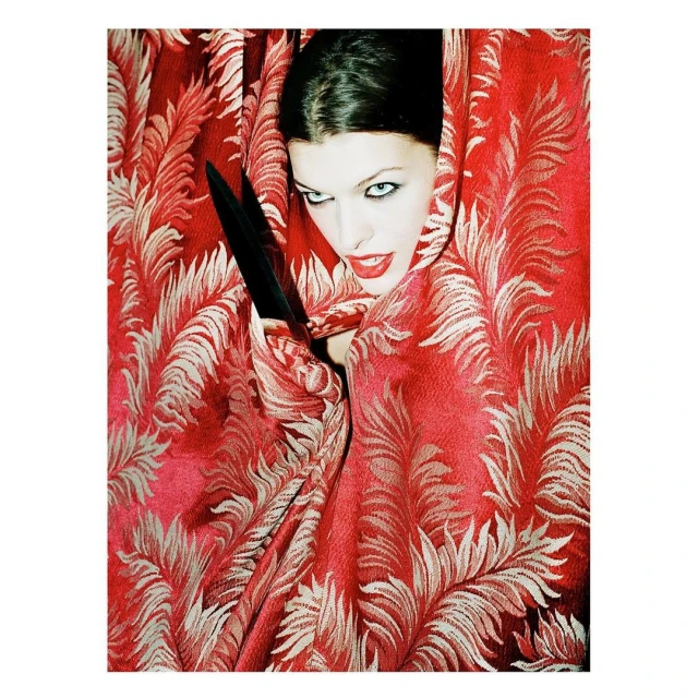 Close-up of a woman with fair skin, dark hair, and red lipstick. She is wrapped in red floral fabric, with a black object in her hand.
