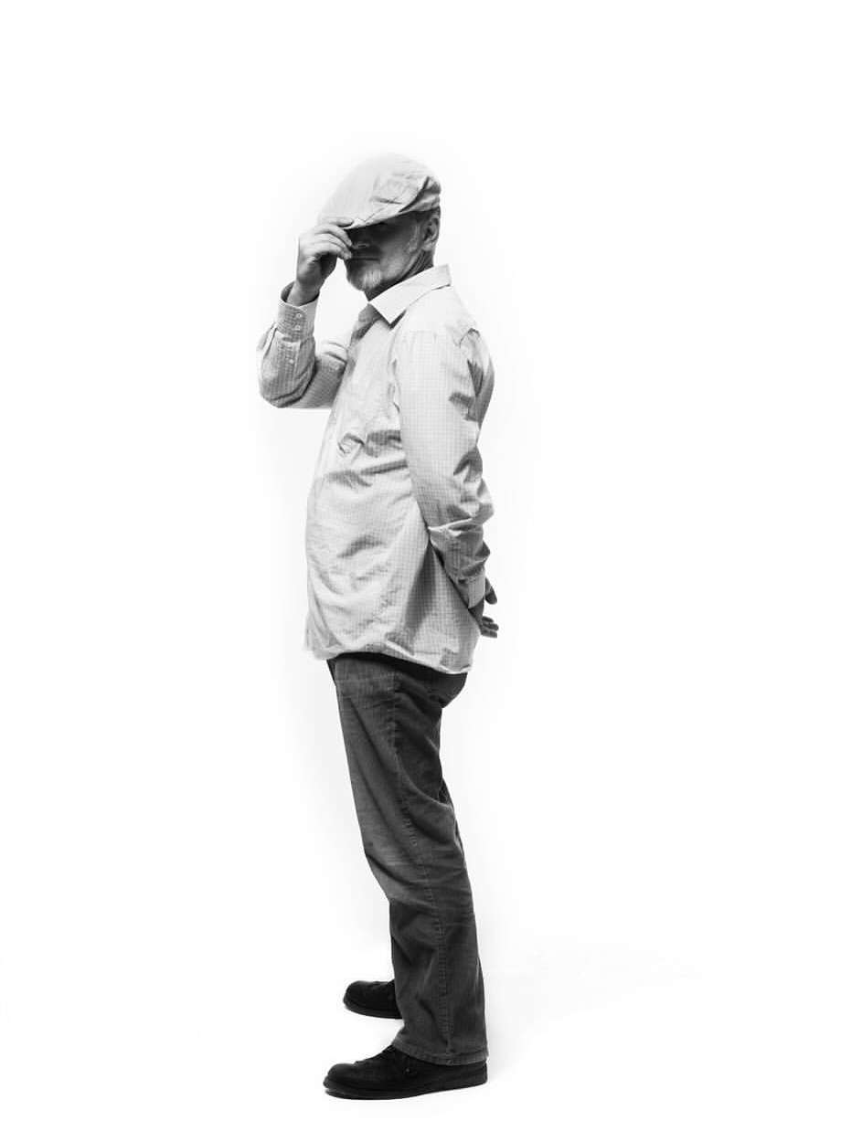 Full-length black and white shot of an elderly man in a newsboy cap, light-colored shirt, dark jeans, and shoes posing on a white background.