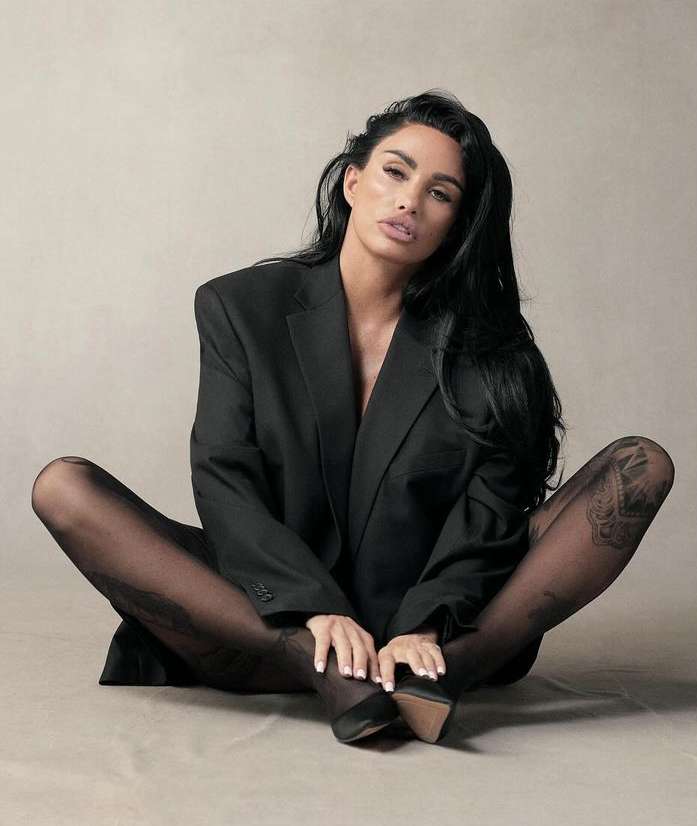 A woman in a black jacket and patterned tights sits on the floor with legs crossed, hands resting on her ankles in a neutral studio.