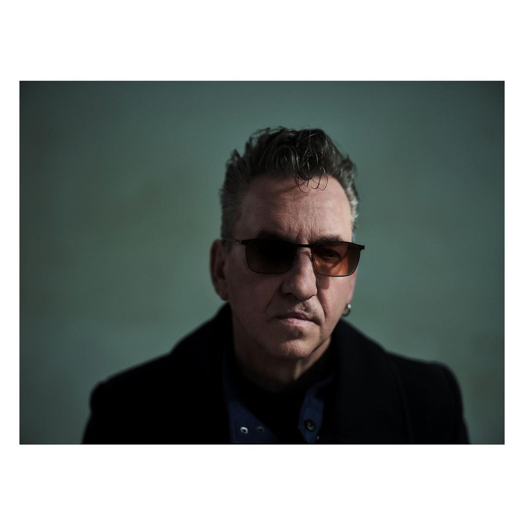 Close-up studio portrait of a mature adult man with dark hair wearing sunglasses and a black coat against a green background.