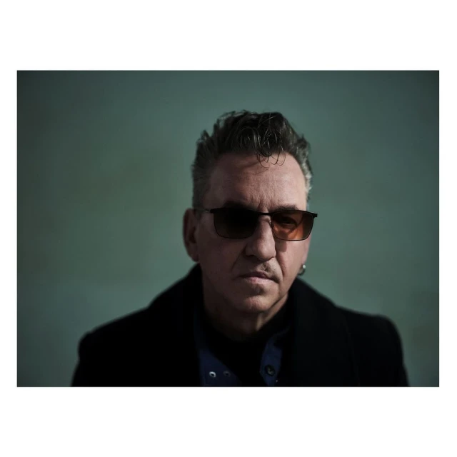 Close-up studio portrait of a mature adult man with dark hair wearing sunglasses and a black coat against a green background.