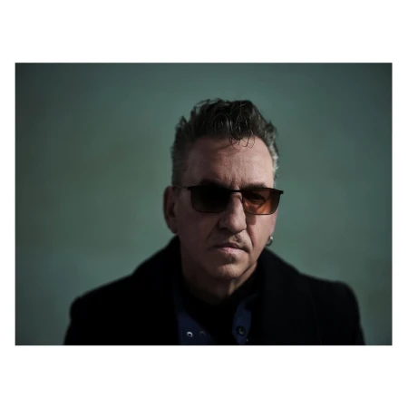 Close-up studio portrait of a mature adult man with dark hair wearing sunglasses and a black coat against a green background.
