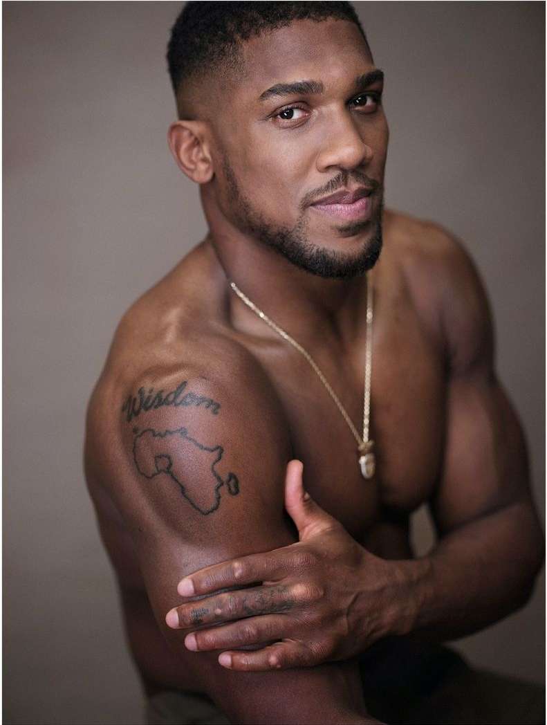 Portrait of a muscular, dark-skinned adult male with a beard and a gold chain, posing shirtless in front of a neutral background.