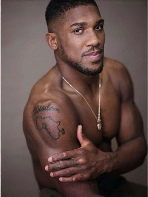 Portrait of a muscular, dark-skinned adult male with a beard and a gold chain, posing shirtless in front of a neutral background.