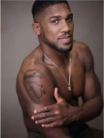Portrait of a muscular, dark-skinned adult male with a beard and a gold chain, posing shirtless in front of a neutral background.