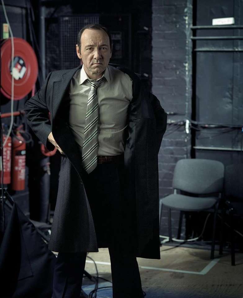 Adult male in suit, tie, and coat standing in a room with brick walls and lighting equipment, looking at the camera with a neutral expression.