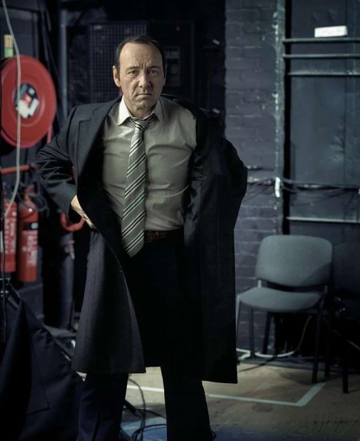 Adult male in suit, tie, and coat standing in a room with brick walls and lighting equipment, looking at the camera with a neutral expression.