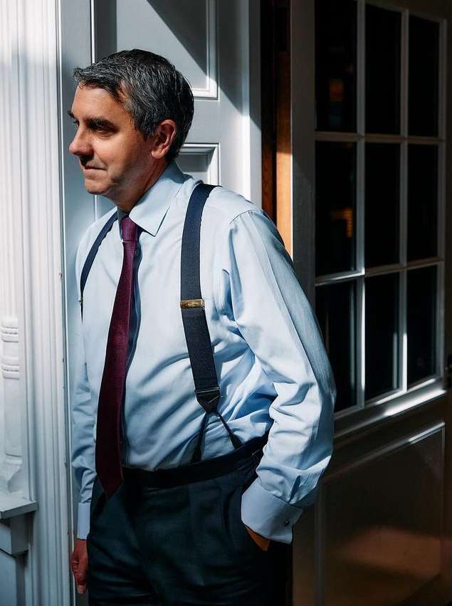 A man in a light blue shirt, suspenders, and dark pants stands indoors near a white door with window panes in a side view, bathed in sunlight.
