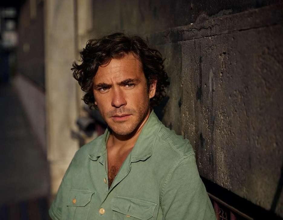 Portrait of a man with dark curly hair wearing a light green button-down shirt, leaning against a dark concrete wall outdoors.