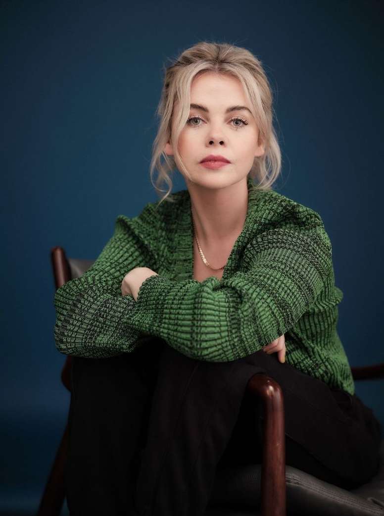 Portrait of a blonde woman seated in a wooden chair wearing a green ribbed knit cardigan against a dark blue background.