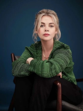 Portrait of a blonde woman seated in a wooden chair wearing a green ribbed knit cardigan against a dark blue background.