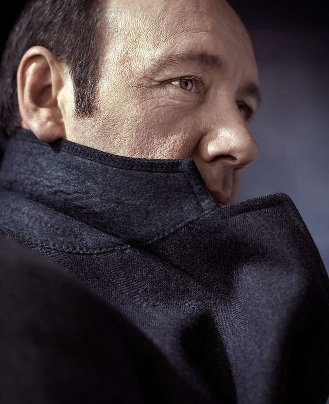 Close-up portrait of an adult male with a neutral expression, wearing a dark overcoat, shot in a studio with controlled lighting.