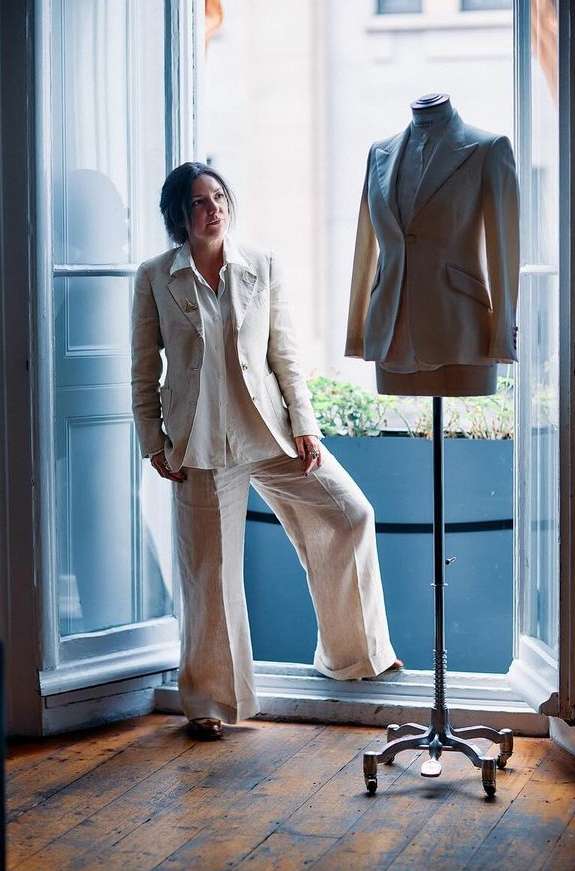 Woman in a linen suit stands in a doorway next to a mannequin in a matching suit, bathed in natural light on a wooden floor.