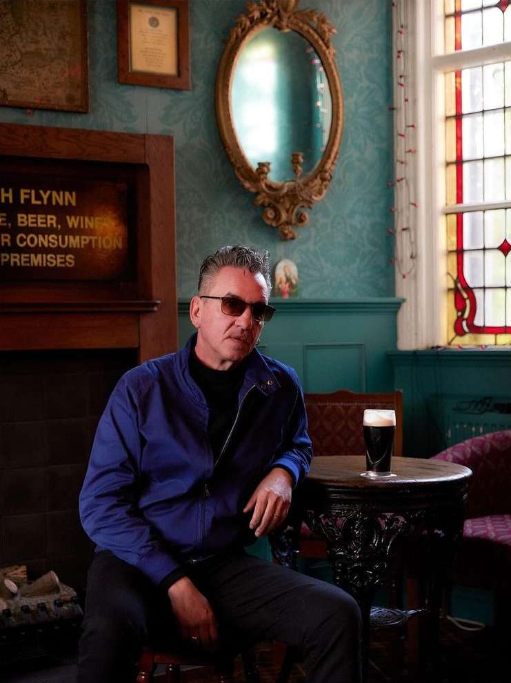 Adult man in sunglasses sits near a dark beer in a pub with decorative walls, a mirror, and window with stained glass.