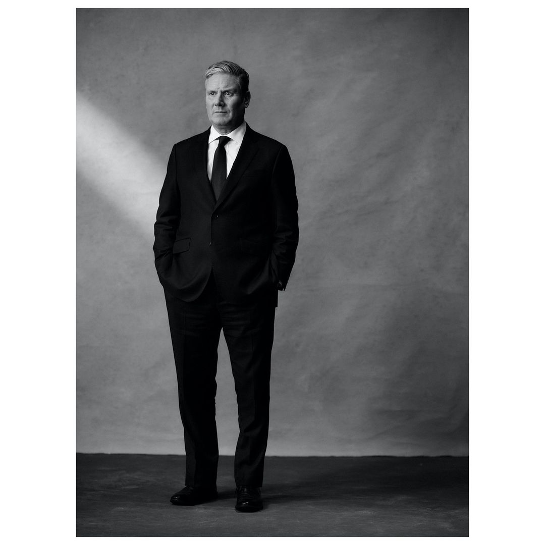 Full portrait of an adult male in a dark suit and tie stands in front of a textured gray background with soft studio lighting.