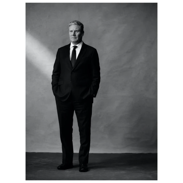 Full portrait of an adult male in a dark suit and tie stands in front of a textured gray background with soft studio lighting.