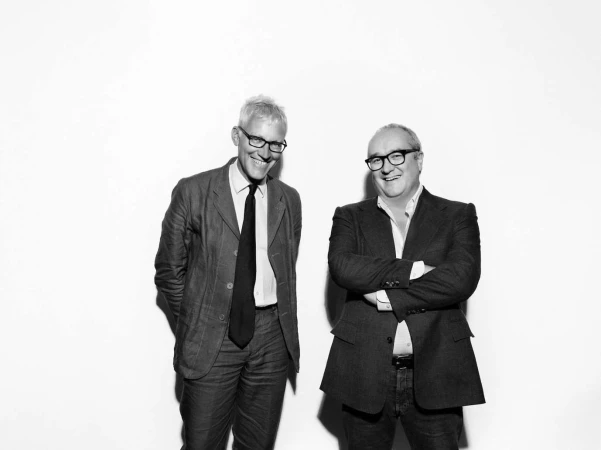 Two men in smart casual suits and glasses smiling against a stark white background in a black and white portrait.