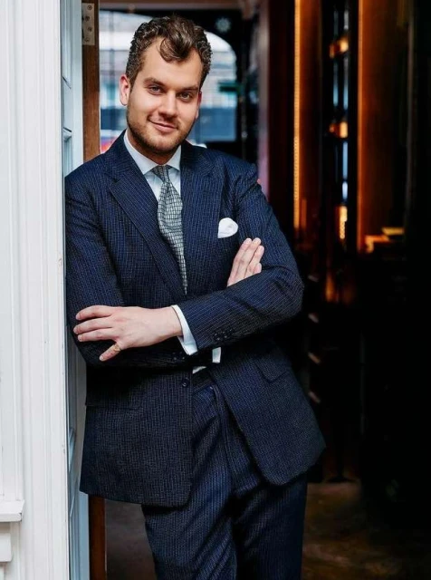 Man in a navy pinstripe suit stands indoors against a white wall with arms crossed, looking at the camera with a slight smile.