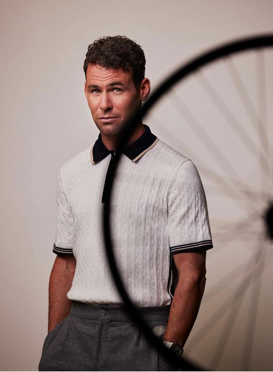 Man in a polo shirt poses in front of a bicycle wheel, hands in pockets, gray background, looking at the camera.