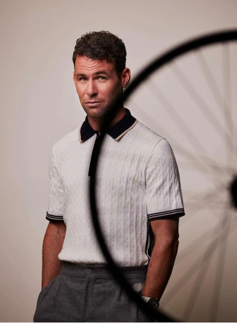 Man in a polo shirt poses in front of a bicycle wheel, hands in pockets, gray background, looking at the camera. 
