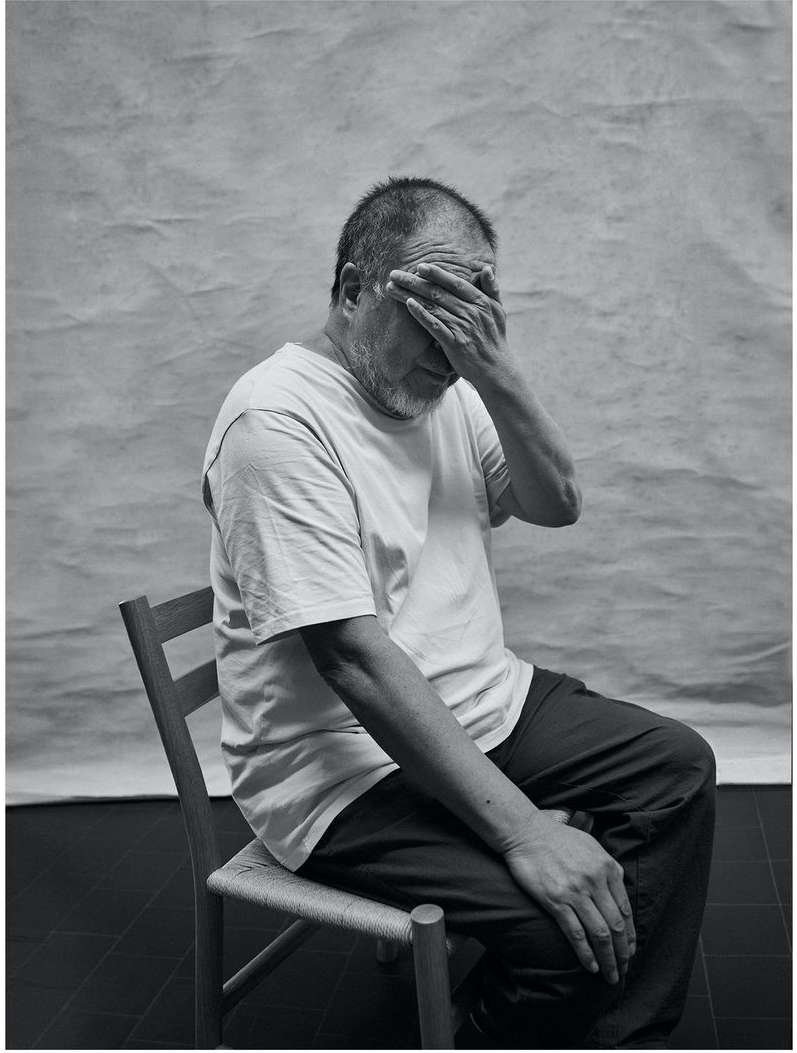 Portrait of an older man wearing a white t-shirt, seated on a chair with his hand covering his eyes against a grey textured backdrop.