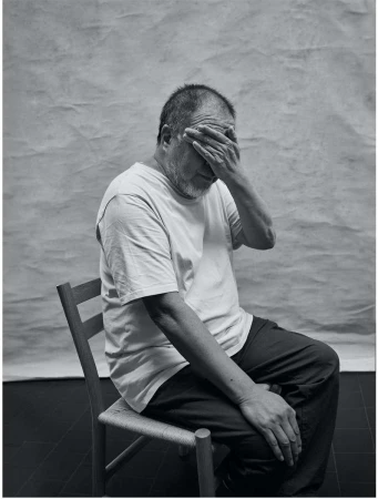 Portrait of an older man wearing a white t-shirt, seated on a chair with his hand covering his eyes against a grey textured backdrop.