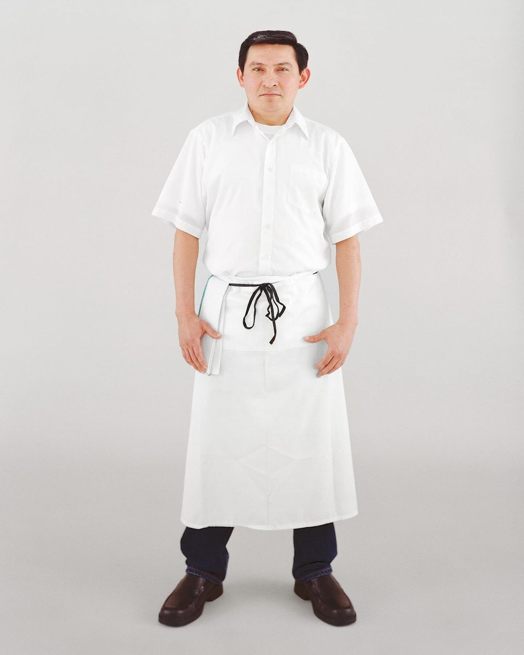 Full shot of a man in a white chef's uniform and apron, hands in pockets, posing against a light gray background in a studio setting.