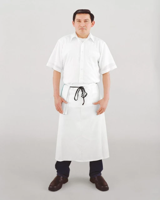 Full shot of a man in a white chef's uniform and apron, hands in pockets, posing against a light gray background in a studio setting.