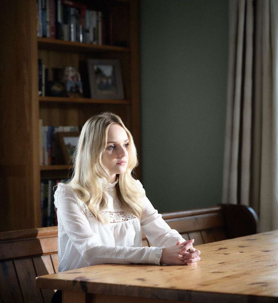 Blond woman with folded hands at a wooden table, bookshelf and window in the background, creating a contemplative indoor portrait.