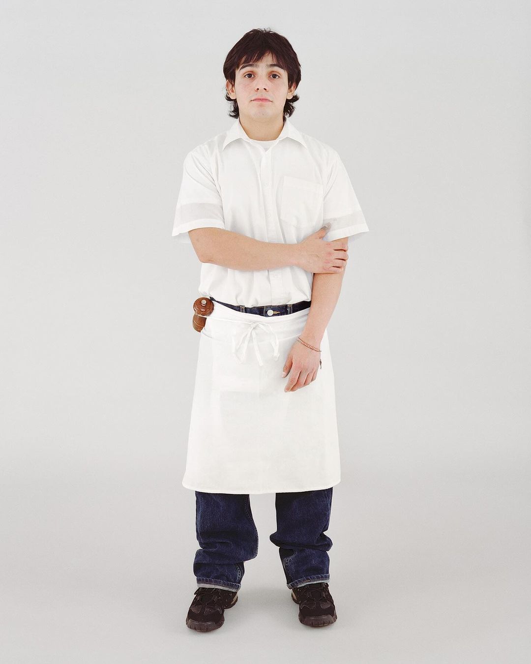 Full-shot studio portrait of a young adult male in a white cook's shirt and apron, dark jeans, with a neutral expression.