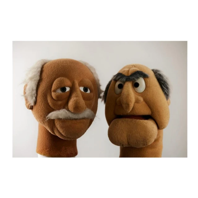 Close-up studio shot of two brown hand puppets with white and black hair, set against a plain white background. Puppets show different expressions.