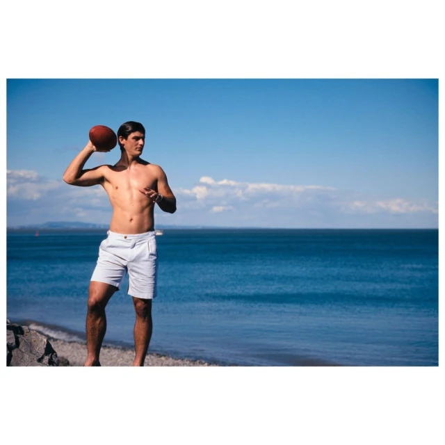 A shirtless man in white shorts throws a football on a sunny beach. Blue sky, ocean, and a rocky shore complete the scene, full body.