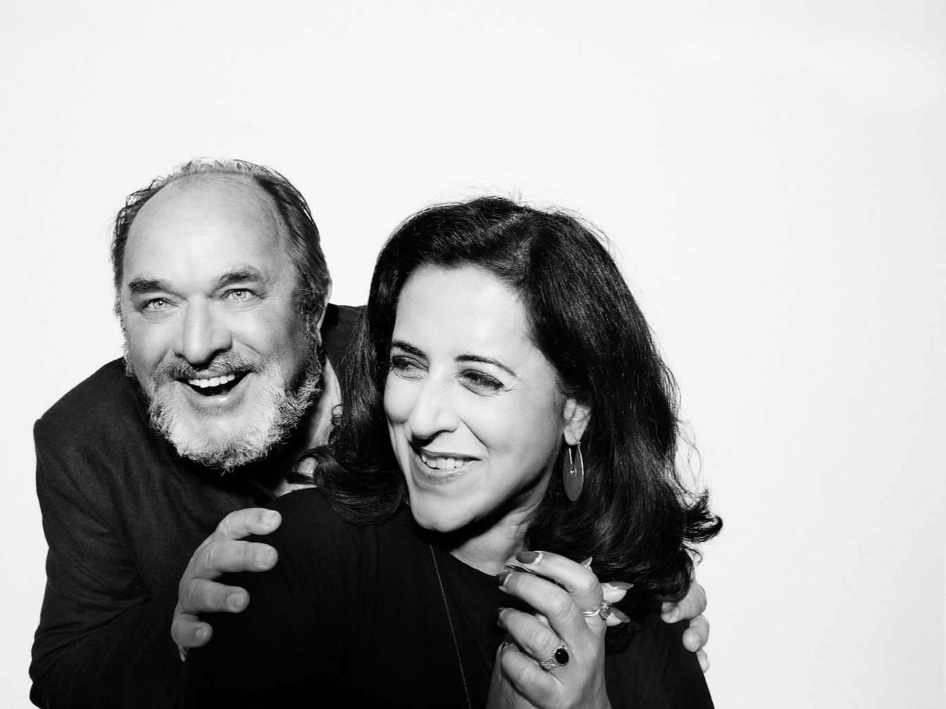 Black and white portrait of a smiling adult man with a beard embracing a smiling adult woman in front of a white background.