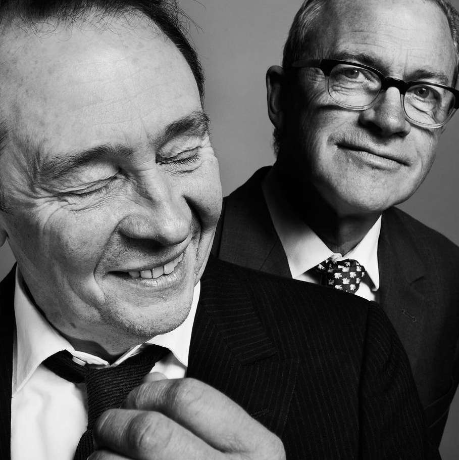 Black and white portrait of two adult men in suits. One man is smiling with his eyes closed, the other stands behind him wearing glasses.