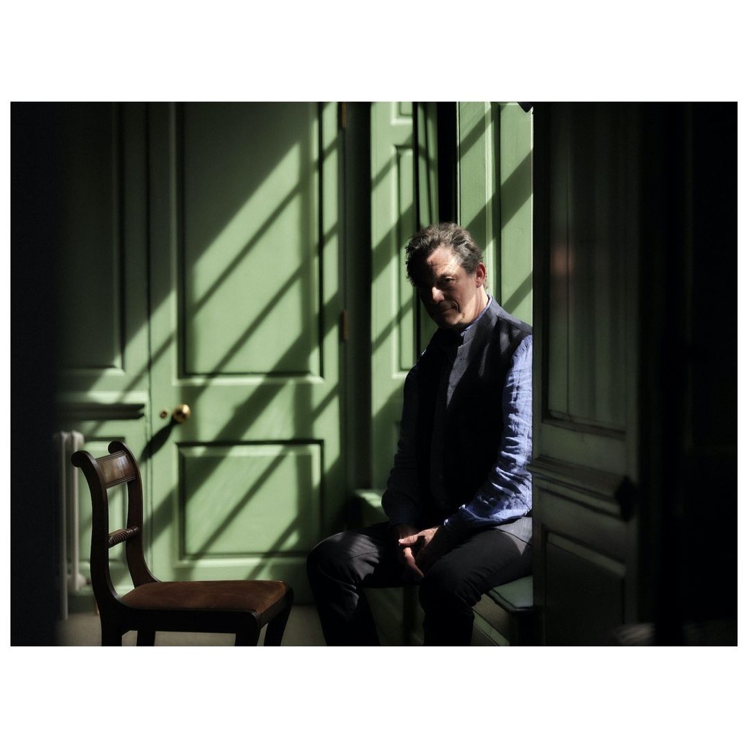 Adult man in a blue shirt and vest sits indoors near a green paneled door with sunlit stripes, next to a brown wooden chair.