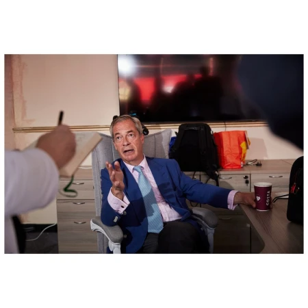 Nigel Farage seated in an office chair wearing a blue blazer and light blue tie while gesturing during an interview in London.