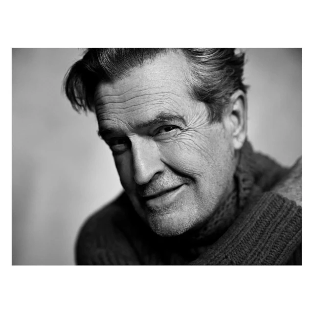 Black and white portrait of an adult male with graying hair, wearing a sweater and smiling softly, captured in a close-up shot indoors.